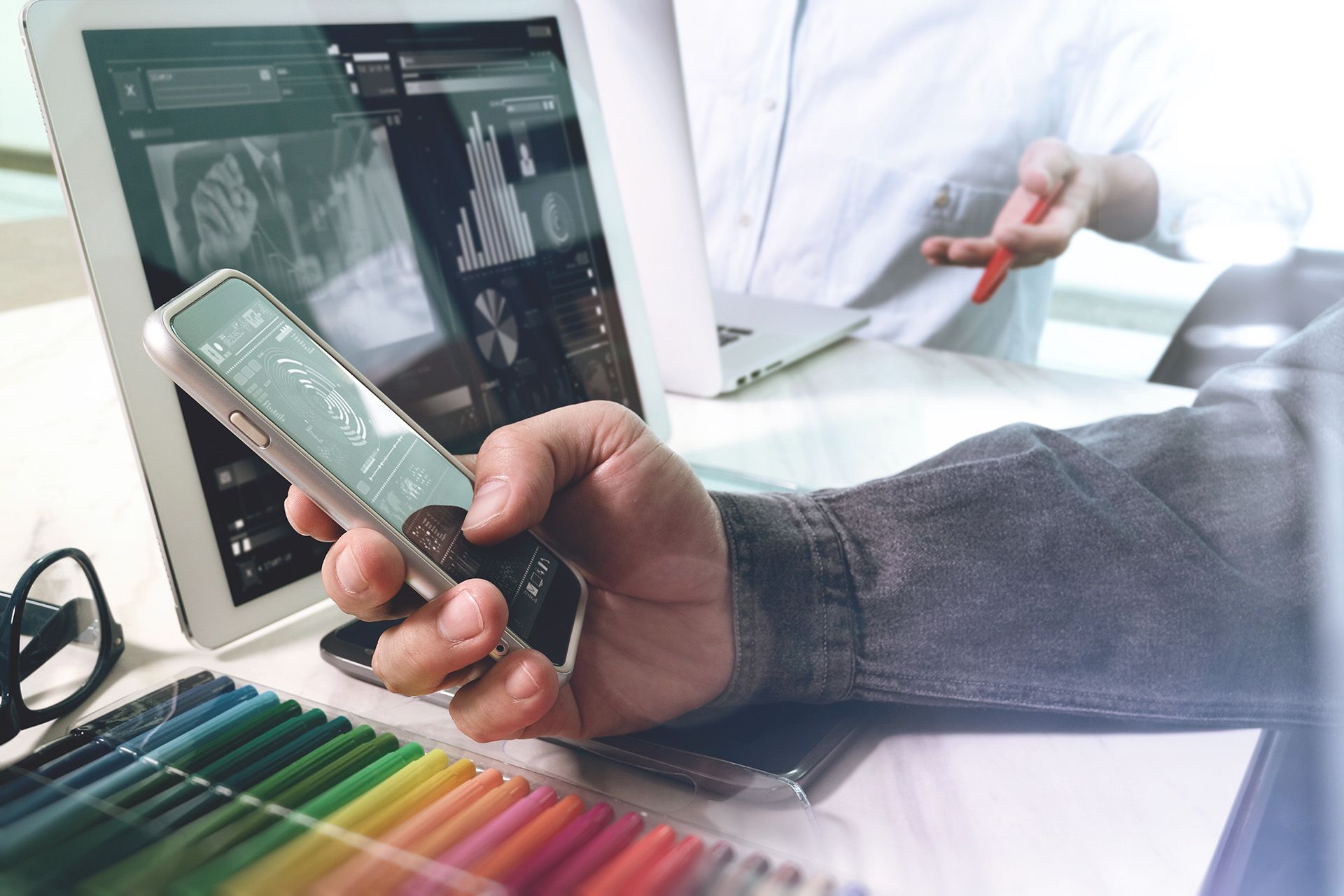 Eine Hand hält ein Handy vor einem Laptop und über Buntstiften in Nahaufnahme